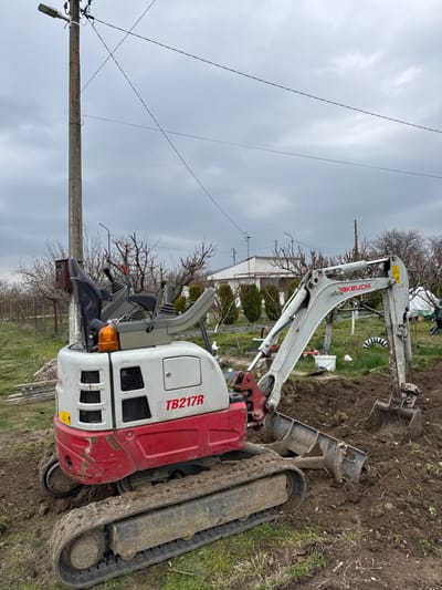 An outdoor scene in Polkovnik Lambrinovo, Bulgaria, depicts a white and red Takeuchi TB217R mini excavator positioned on disturbed earth under an overcast sky. The excavator, facing right, has its tracks covered in dirt, and its bucket is partially embedded in a mound of freshly dug soil, suggesting ongoing excavation. The operator's seat, controls, and an amber warning beacon are visible on the machine. To the left of the excavator, a concrete utility pole equipped with a street light and multiple overhead power lines stands vertically. Bare deciduous trees and shrubs are scattered across the midground, with a wire fence extending from the left. In the background, a white building with dark windows is visible, fronted by a line of evergreen trees. No individuals are present in the frame.