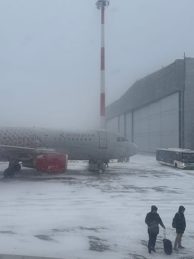 The image displays an airport apron at Emelyanovo Airport, Russia, under conditions of heavy snowfall and low visibility. A grey Rossiya Airlines aircraft, identified by "ROSSIYA" on its fuselage and a geometric pattern on the rear, is centrally positioned on the snow-covered tarmac. The port engine cowling is red. Ground service equipment is visible near the aircraft's nose landing gear. A tall red and white striped mast rises vertically behind the aircraft. To the right, a large hangar building with multiple light-colored bay doors is partially visible through the falling snow. A dark airport bus, with "COLLS" faintly visible on its side, is parked near the hangar. In the foreground, two individuals, clad in dark winter coats with hoods, walk on the snow-covered apron from right to left. One individual pulls a dark roller suitcase, and the other carries a backpack. Snowflakes are evident across the entire scene.