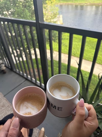The image captures two hands holding coffee mugs, likely on a patio or balcony. One mug is white with the text "HOME", and the other is pink. The mugs appear to contain lattes, each with a creamy foam swirl on top. Through a screen or railing, a view of a green lawn, a paved walkway, and some trees is visible. Given the prevalence of lush greenery, walkways and screened patios, the location may be a residential area with similar aesthetics in Orlando, United States. The person in the image is possibly enjoying a peaceful moment with a drink, likely in the morning or early afternoon, as implied by the outdoor lighting. The overall atmosphere seems relaxed and domestic.