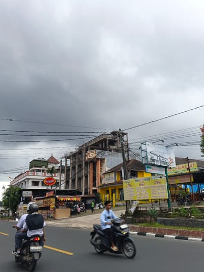The image captures a street scene under an overcast sky, showing commercial activity and construction. The signs and language visible, particularly Indonesian text like "Menu Best Seller" listing dishes such as "Sate Usus Pari," "Ayam Bakar Madu," and "Nasi Goreng Kampung," along with the Indonesian license plate format (e.g., F 2416 WX), indicate the location is in Indonesia, not Palayan, Philippines.
In the foreground, an asphalt road features a solid yellow line separating lanes. Two motorcycles are in motion from right to left. The motorcycle further left carries two individuals; the driver wears a white shirt and helmet, while the passenger, also in a white shirt, wears a black backpack. The second motorcycle, to the right, is ridden by a solo male in a light blue collared shirt, without a helmet.
The background consists of multiple structures. On the left, a multi-story building with white and red sections features a distinctive green-domed roof element and a visible "Lazzato" sign. Adjacent to it, a multi-story concrete building is under construction, with exposed rebar and scaffolding-like elements. To the right of the construction, a series of single and two-story commercial buildings are visible, painted predominantly yellow. One building displays a large "Menu Best Seller" board with Indonesian food items. Further right, a sign for "Raja Laut" (Seafood King) is present. Overhead, numerous electrical and utility lines crisscross the frame. The immediate roadside features a low wall and some sparse vegetation.