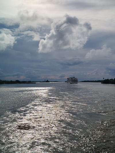 A wide, choppy river flows under a dramatic sky filled with dark, heavy clouds and bright patches of sunlight. A large, multi-decked white boat is the prominent subject, traveling away from the viewer toward the right side of the frame. To the left, a smaller barge with a white cabin is visible, also moving away. The river's surface shimmers with sunlight, creating a strong reflection. The distant shoreline is lined with dark green trees and some low buildings, with a lighthouse-like structure visible on the horizon. The scene suggests a possibly stormy or overcast day with intermittent sun. The location is identified as Kargulino, Russia.