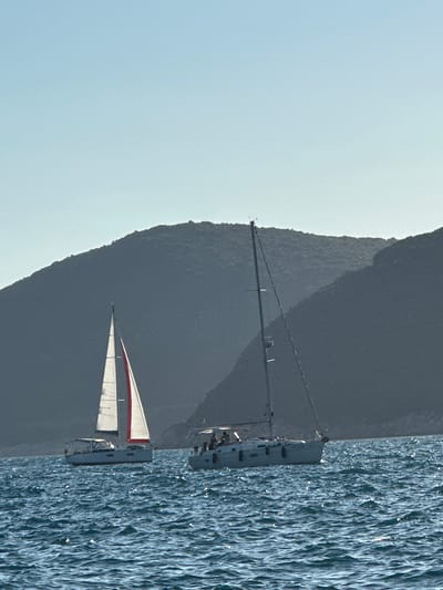 The image captures a maritime scene near Luštica, Montenegro, under a clear, light blue sky. Two monohull sailboats are visible on choppy, dark blue water reflecting sunlight.
On the left, a white sailboat, smaller than the one on the right, navigates with its main sail and jib partially unfurled. The jib displays a white fabric with a prominent vertical red stripe. The boat features a soft-top bimini or dodger and a series of dark windows along its hull. A wind indicator is visible at the top of its mast. No occupants are clearly discernible on this vessel.
To the right, a larger white sailboat is depicted without its sails unfurled. Its tall mast is equipped with an anemometer and other instruments at its apex, and multiple spreaders are visible along its length. At least five individuals are present on the boat's deck, primarily in the cockpit area and midship, with their specific actions not clearly defined beyond being aboard. Several dark, cylindrical fenders are visible hanging along the starboard side of the hull.
In the background, a series of dark, heavily forested hills or mountains rise, creating a silhouette against the sky. The undulating terrain suggests a rugged coastline. The lighting indicates sunlight from the right, creating dappled reflections on the water's surface.