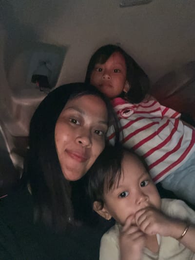 This is a medium shot, horizontally oriented, low-angle, candid photograph of a woman and two children lying down. The subjects appear to be in a bed, as indicated by the presence of bedding which includes a blanket with red and white stripes and a white pillow. The lighting is dim, suggesting it might be nighttime or a dimly lit room.
The woman is in the foreground, with her face close to the camera, looking upwards. She has dark hair and appears to be smiling gently. Next to her, to her right, is a baby or young toddler who is looking at the camera with wide, dark eyes. The child has its hand near its mouth. To the woman's left is another child, possibly older, with dark hair. This child is also looking towards the camera, with eyes open and a slight smile. All three subjects are nestled together, suggesting a moment of intimacy or relaxation.
The overall mood of the photograph is warm and familial. The dim lighting and close proximity of the subjects contribute to a sense of coziness. There are no visible objects other than the bedding and the people. There is no discernible text in the image. The location context provided is Ngadirejo, Indonesia.