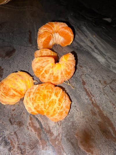 Four peeled citrus fruits, consistent with tangerines or mandarins, are depicted resting on a dark, mottled surface. The surface displays variations in color, ranging from deep charcoal grey to black, with discernible lighter grey streaks and some reddish-brown marbling or staining. One fruit, located at the top center, is partially separated into segments. Another fully peeled fruit is positioned centrally towards the bottom. A third fully peeled fruit lies in the bottom-left quadrant. A smaller cluster of peeled segments is situated between the upper and lower central fruits. Minimal white pith adheres to the fruit surfaces. The scene is located in Saint-Étienne, France.