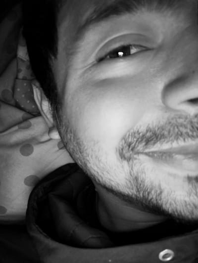 This black and white, close-up photograph features a person, likely male, with facial hair. The composition focuses tightly on the right side of the individual's face (viewer's left), encompassing one eye, part of the cheek, and the mouth/chin area.

The subject is looking directly towards the camera with a subtle, possibly smirking or soft smiling expression. The right eye is open, and a distinct bright reflection, a catchlight, is visible in the pupil, indicating a light source. Fine lines are apparent around the eye, suggesting a dynamic expression. A light beard or prominent stubble covers the cheek, jawline, and area around the mouth. The mouth is slightly open, revealing a glimpse of teeth.

Behind the person's head, to the left of the frame, there is a soft, patterned fabric with light-colored polka dots on a darker background, possibly a pillowcase or blanket, suggesting an indoor setting. At the bottom of the image, dark, possibly padded clothing, like a jacket or coat collar, is visible, featuring a metal eyelet on the lower right edge. The overall impression is an intimate, perhaps self-taken, portrait. No text is visible in the image. The time of day and weather conditions are not discernible from the photograph. The provided location context is Moaña, Spain.