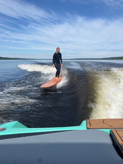 In Gromovo, Russia, on a bright, clear day with a sky full of wispy clouds, a woman wearing a black wetsuit surfs on a wakeboard on a dark body of water. The woman stands on an orange wakeboard, her body angled slightly towards the left of the frame. She appears to be balanced and in control, her hair tied back. Behind her, a large wave is being created by a boat, which is partially visible in the foreground. The boat is teal and gray, with brown accents on the deck. The water is dark and choppy, with white foam from the wake. In the distance, a tree-lined shore is visible under the vast blue sky. The scene captures a moment of active recreation on the water.