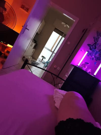 The image depicts an interior scene, possibly a bedroom, bathed in the warm glow of purple and orange lighting. In the foreground, a pink duvet covers a bed, and a portion of a person's leg, clad in black lace, is visible. To the left, a large black fan is positioned near a television set, casting an orange light on the wall and a framed picture. A white door stands ajar, revealing a hallway with white cabinets and a tiled floor. A window with vertical blinds allows natural light to enter the room.
On the right side of the image, purple LED lights illuminate a black cabinet adorned with various butterfly decals and a small decorative fishbowl containing figures. The room appears to be a personal space, possibly within a residential apartment or house. No specific landmarks or indications point to the city of Komotini, Greece.