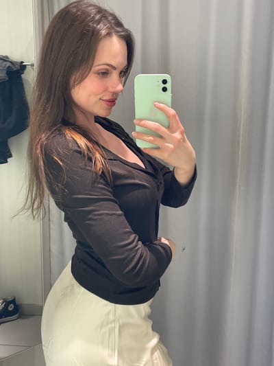 A female subject with long, dark brown hair and fair complexion is depicted from the waist up in what appears to be a modern retail changing room. She is wearing a dark, long-sleeved top with a v-neck and a light-colored, possibly cream or white, skirt. Her right hand holds a light green smartphone with two visible rear camera lenses, positioned to capture her reflection in an unseen mirror. She is looking towards the phone's screen. The background consists of plain grey walls or curtains. To the immediate left, a metal clothing rail is visible, from which a dark garment hangs, and a portion of a black high-top sneaker rests on a surface below the rail. The image does not depict any location within the city of Pompei, Italy.