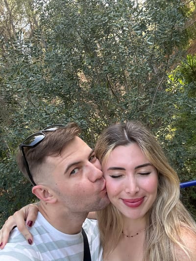 A man and woman are photographed together, possibly in Altea, Spain, based on the typical Mediterranean vegetation in the background. The woman has blonde hair and is wearing a necklace; her eyes are closed and she is smiling. The man has short brown hair and is wearing sunglasses on the back of his head; he is kissing the woman's face. They are positioned in front of a dense background of green foliage. The woman's hand is visible, showing red nail polish. A white and green striped shirt is visible on at least one of them. The photo appears to be taken outdoors in daylight.