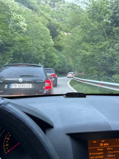 The image, taken from inside a vehicle in what appears to be heavy traffic near Бачковски манастир, Bulgaria, shows the dashboard, steering wheel, and the road ahead through the windshield. The dashboard display reads "Ot utre," with a range of 930 km and a consumption rate of 3.5 Ltr/100 km. A black car with the license plate "PB 3242 CE" is visible in front, followed by at least two more vehicles. A guardrail borders the road, and a dense forest lines the roadside.