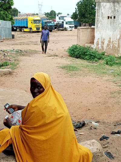 In Gadao, Nigeria, a person wearing a blue t-shirt and dark pants walks across a dusty, open area. In the background, several trucks are parked, including a bright yellow truck with a blue trailer and a white truck. To the left, a corrugated metal structure is visible. In the foreground, a person wrapped in a vibrant yellow headscarf and garment is seated, holding a black mobile phone and what appears to be a small white card in their hands. The ground is unpaved and scattered with debris, including plastic bags and other refuse. A textured, light-colored building with dark rectangular openings is visible to the right. Green foliage grows in patches around the building and along the ground.