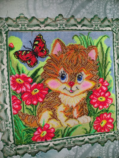 A beaded embroidery of a kitten and a butterfly is displayed in a decorative green and white frame. The kitten, depicted with orange and brown fur, blue eyes, and pink cheeks, sits amidst red flowers and green grass. A red and black butterfly hovers in the upper left corner. The background is a light blue sky. The artwork is likely a craft project, possibly intended for home decoration. The location is the village of Shpychyntsi, Ukraine.