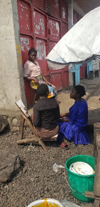 The scene depicts an outdoor setting in Buzove, Democratic Republic of the Congo. Three females are present: one stands leaning against a weathered red metal storefront door, wearing a light-colored, polka-dotted top and a patterned skirt; another is seated on a makeshift wooden stool with her back partially to the viewer, wearing a dark top and light brown bottom; the third is seated on the ground facing the stool, clad in a vibrant blue dress with red sandals visible near her feet. The two seated individuals appear to be conversing. Overhead, a large, worn white market umbrella provides shade. The ground consists of dark gravel and small stones. To the left, a concrete pillar is visible. In the foreground, a green bucket holds several white dishes, and a white bowl containing a yellow-orange substance rests on the ground.