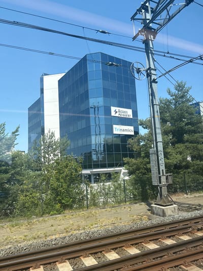 This media file depicts a view from a train window, showcasing an urban environment in Eindhoven, Netherlands. The primary subjects include a modern glass-fronted office building, overhead power lines typical of a train infrastructure, and a section of railway tracks in the foreground.
The office building features a striking facade of blue-tinted glass panels. Two prominent signs are visible on the building's exterior. One displays the text "Komers Advocaten" with a triangular logo. The other sign reads "Trinamics" in blue lettering. The building appears to be several stories high, with a white section visible on its lower floors.
Adjacent to the building, a network of overhead power lines and support structures for electric trains are evident, indicating that the viewer is likely on a moving train. The railway tracks, complete with ballast and sleepers, run diagonally across the right side of the frame, further confirming the setting.
The scene is illuminated by bright sunlight, suggesting a clear day, likely during daytime hours. Green trees and foliage are interspersed between the building and the railway, adding a touch of nature to the urban landscape. The overall impression is one of an active, modern city with well-developed transportation infrastructure. No people are directly visible, and the image focuses on the architectural and infrastructural elements of the scene. The graffiti on the overhead structure is a small detail that adds a touch of urban authenticity.