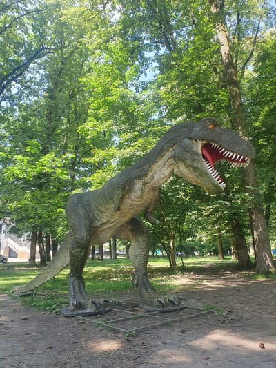 The image shows a life-size statue of a Tyrannosaurus Rex dinosaur standing in a park-like setting. The dinosaur is depicted with its mouth open, revealing sharp teeth and a red interior. It is positioned on a flat, square base with metal supports. The surrounding environment consists of lush green trees with dappled sunlight filtering through the leaves, creating shadows on the dirt path and grass. In the background, there are more trees, a pathway, and a glimpse of a building with stairs and a parked car. The scene appears to be outdoors during the daytime, under a clear sky. This statue is located at the dinosaur park "Fabulous Dino Land" in Ivano-Frankivsk, Ukraine.