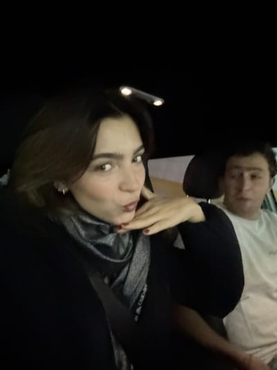 A brunette woman poses for a selfie with pouty lips, wearing a black jacket and a metallic gray top. Her hand is casually placed near her chin. Behind her, a man in a white t-shirt rests his head on what appears to be a seat or headrest. The lighting is dim, casting the background into darkness.