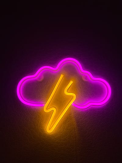 A neon sign shaped like a cloud with a lightning bolt inside is mounted on a textured dark wall. The cloud is outlined in pink neon, and the lightning bolt is in yellow neon. The sign is illuminated, casting a glow on the wall. The background is dark and textured, with subtle lighting effects from the neon sign. There are no people or other objects visible in the image. The location of Ильинское-Усово, Russia, is not discernible from the image itself.