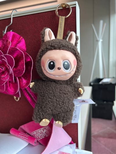 This image depicts an indoor scene within the city of Garston, United Kingdom. The central subject is a brown, textured plush keychain toy, identifiable by its tag bearing "THE MONSTERS." This toy features a light-toned face with large black eyes, a small nose, and a wide, smiling mouth revealing small white teeth. It is encased in a dark brown, shaggy suit with prominent light brown-lined ears, suggestive of a rabbit or similar animal. Small, pale hands and feet protrude from the suit.
The plush toy hangs from a golden keyring attached to a beige fabric strap, also labeled "THE MONSTERS," which is suspended from the top edge of a partition. To the left of the plush toy, a large, metallic fuchsia-colored decorative item, shaped like a multi-layered flower or rosette, is partially visible and appears to be secured by a silver S-hook at the upper left.
The immediate background for these items is a dark red, textured surface, resembling a fabric-covered bulletin board or partition.
In the foreground, below the plush toy, a collection of items is loosely arranged. These include a bright pink ribbon adorned with multiple rows of silver rhinestones, a plain pink grosgrain ribbon, and crumpled white papers or fabric.
The background to the right is out of focus, revealing a broader indoor environment. A light-colored object with multiple thin, upward-pointing arms, possibly a modern coat rack or abstract sculpture, stands further back. Beyond this, two dark-colored, rectangular objects, possibly storage containers or furniture, are stacked. The floor in this background area is light-colored and exhibits a subtle geometric pattern, suggesting a rug or specific tiling. Ambient light enters from the right side, illuminating the scene. No individuals are present, and all objects are stationary.