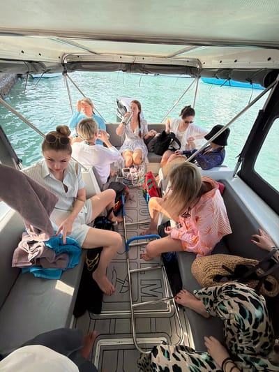 The image depicts a group of eight women inside a boat on the water, presumably in the city of Maafushi, Maldives. The women are seated on upholstered benches along the sides of the boat, which is partially covered by a canopy. The water is visible outside the boat, suggesting they are traveling on it. The women are dressed in casual clothing suitable for a vacation setting, including shorts, dresses, and swimsuits, indicating a relaxed and informal atmosphere. Some of the women are barefoot. Objects such as beach bags, water bottles, and towels are scattered throughout the boat, suggesting they are prepared for activities such as swimming or sunbathing. The floor of the boat has a gray patterned surface with the word "SHADOW" printed on it. The overall scene suggests a leisurely boat trip with a group of friends or acquaintances enjoying a vacation in Maafushi.