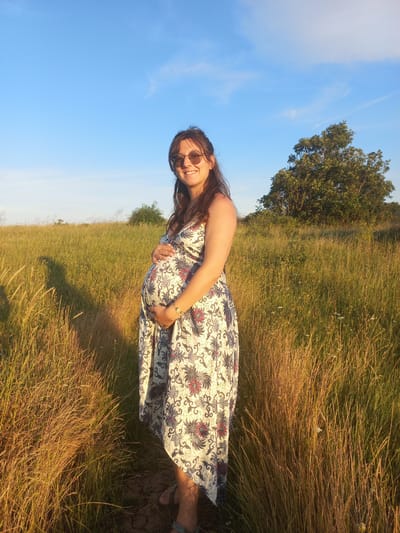 A pregnant woman stands in a sunlit field of tall grass, her hands resting on her belly. She is wearing a patterned dress and sunglasses, and a tree is visible in the background. The sky above is a clear blue with a few wispy clouds. The scene appears to be set in a natural landscape, possibly in the vicinity of Sinemorets, Bulgaria, given the dense vegetation and the golden hour lighting that suggests a late afternoon or early evening.