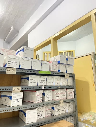 The image depicts a storage area, likely a pharmacy or medical supply room, in Margoyoso, Indonesia. The primary subjects are numerous boxes of medication neatly stacked on metal shelves. The boxes are predominantly white with blue and red accents, and many have handwritten labels with dates and names, indicating inventory management. Some labels clearly show "ED" followed by dates like "7/26", "10/26", and "12/26", signifying expiration dates.
The scene is indoors, with a brightly lit ceiling and walls. In the background, a yellow wall and a window with horizontal blinds are visible, suggesting a functional but not necessarily modern setting. To the left, a white wall with a pipe is present. The lighting suggests it could be daytime.
The activity taking place appears to be the organization and storage of medical supplies. The meticulous arrangement of the boxes implies a system for tracking stock and ensuring timely use of medications before their expiration. The handwritten notes suggest manual inventory control, possibly by healthcare workers.
Visible text includes "OBAT PROGRAM PEMERINTAH HIBAH" (Government Sponsored Medicine Donation), "MINUM OBAT SECARA TERATUR DAN TUNTAS WAN SEMBUH" (Take medicine regularly and completely to get well), and various names and dates that appear to be inventory records. This text provides context about the nature of the supplies and their intended use, highlighting a focus on public health initiatives. The overall impression is one of an organized, functional space dedicated to managing essential medical supplies.