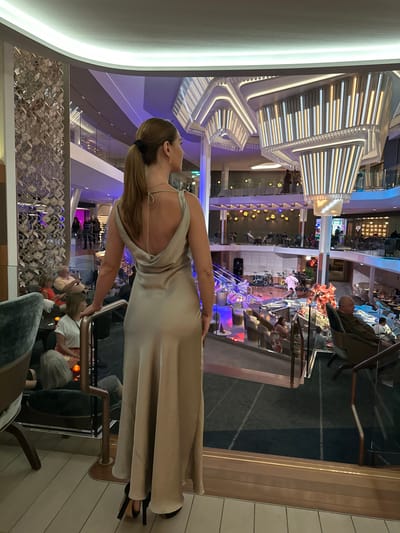 An interior scene captured in the city of Punta de Maisi, Cuba, depicts a female subject from behind, standing on an elevated observation deck overlooking a multi-level atrium. The subject, with light brown hair pulled into a high ponytail, wears a full-length, champagne-colored, backless satin gown with a cowl neckline and black high-heeled shoes. Her right hand rests on a copper-toned railing that separates the platform from the expansive space below.

The atrium is characterized by its modern, luxurious design and artificial illumination. Overhead, two large, multi-tiered chandeliers with geometric forms and vertical light panels emit bright white and warm light. The space features multiple levels connected by stairs, with glass and copper-toned railings defining each balcony. The background includes various seating arrangements, such as armchairs and small tables, populated by multiple individuals. On a central lower level, a stage is set up with a drum kit, keyboards, and other musical equipment, indicating a performance venue. A person in white attire is visible near the stage. The environment is brightly lit with a blend of white, purple, and blue LED lights, suggesting an evening setting or a controlled indoor atmosphere. A decorative metallic wall panel with a geometric pattern is present on the left side of the upper level. The flooring in the immediate foreground consists of white planks, transitioning to dark carpet on the lower levels. Individuals in the background appear to be socializing, seated, or observing the stage area.