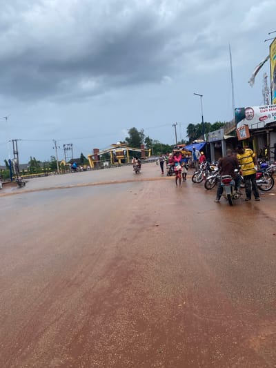The image depicts a street scene near the Auchi Polytechnic in Auchi, Nigeria, under an overcast sky with heavy grey clouds. The reddish-brown road surface appears wet, suggesting recent rain.
In the mid-ground, an archway structure clearly marked "POLYTECHNIC AUCHI" serves as a prominent landmark. To the right of the archway, various commercial structures line the road, some featuring blue tarpaulin awnings. Above these structures, a pedestrian bridge spans the area, and multiple power poles with electrical lines are visible. Further to the right and in the background, tall communication towers are present.
On the right side of the road, several individuals are congregated around multiple motorcycles (okadas). One man in a brown shirt is seated on a motorcycle, while another man in a yellow and black striped shirt stands beside him. A large billboard with a political advertisement displaying a man's portrait and text including "YOUR TRUST," "OUR MAN," and "Vote" is affixed to a building on the right.
Further into the scene, a woman in a pink top and light-colored skirt is walking, carrying goods balanced on her head. Other individuals are scattered across the scene, either walking or riding motorcycles. The general activity indicates a bustling street environment with everyday pedestrian and vehicular movement.