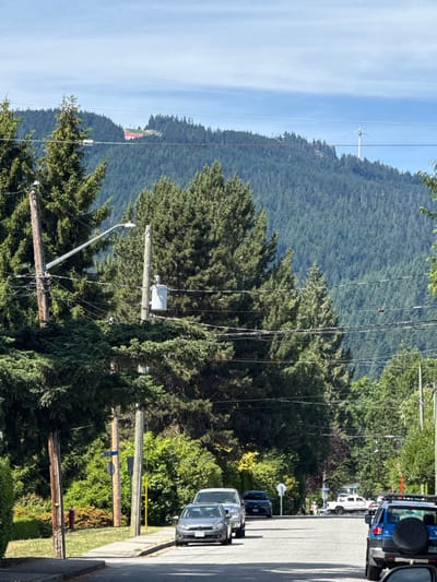 The image is a street-level view looking uphill towards a wooded mountainside in North Vancouver, Canada. In the foreground, a residential street is lined with parked cars and sidewalk. Utility poles and power lines are prominent, stretching across the frame. Lush green trees and bushes are abundant. In the middle ground, the slope of the mountain is covered in dense evergreen forest. In the background, a large red building is visible on the crest of the mountain, with a wind turbine further to the right. The sky is blue with scattered white clouds. The scene appears to be during daylight on a clear day.