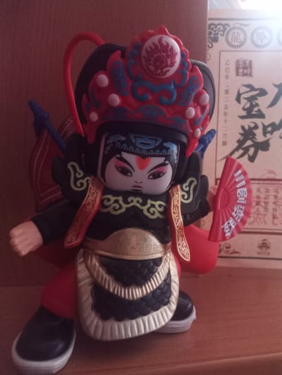 A close-up, slightly blurry, eye-level medium shot captures a doll resembling a Peking Opera character. The doll is positioned on a wooden surface. The doll is adorned with a red and blue headdress, black armor with gold trim, and red leggings. It holds a red fan in its right hand, which has white Chinese characters. The background features a faded, textured wall and a paper item with Chinese calligraphy. The lighting is warm, casting soft shadows. There are no people or animals present in the image, and the doll is the sole subject. There is no indication of the location being Usman, Russia.