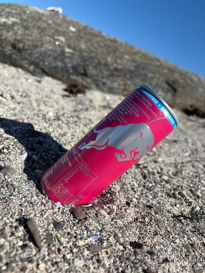 The image displays a Red Bull Peach Edition Sugarfree energy drink can, predominantly pink/red with a silver bull logo, resting on a pebbly and sandy surface. The ground is composed of various small stones, sand, and numerous tiny seashells, strongly suggesting a beach or shoreline in Nærsnes, Norway.
In the background, a large, blurry grey object, possibly a rock formation or driftwood, spans diagonally across the left side of the frame. Above this object and in the distance, a clear, bright blue sky is visible, indicating sunny weather. The bright light casts subtle shadows on the ground, suggesting it is daytime, possibly morning or late afternoon.
No activity or event is actively taking place; the can appears to be discarded or placed on the ground.
Visible text on the can includes "SUGARFREE" on the blue top rim, and "Peach Edition Sugarfree." Further details are in what appears to be Norwegian, such as "redusere tretthet og utmattelse" (reduce tiredness and fatigue), "smak av hvit fersken" (taste of white peach), and "Høyt koffeininnhold" (high caffeine content), along with warnings for "gravide" (pregnant women). Nutritional information per 100 ml is partially legible, including "12 kJ (3 kcal)" and percentages of daily intake for vitamins: Niacin (6.4 mg / 40%), Vitamin B6 (2 mg / 33%), Folic acid (0.2 mg / 14%), and Vitamin B12 (0.4 µg / 16%).