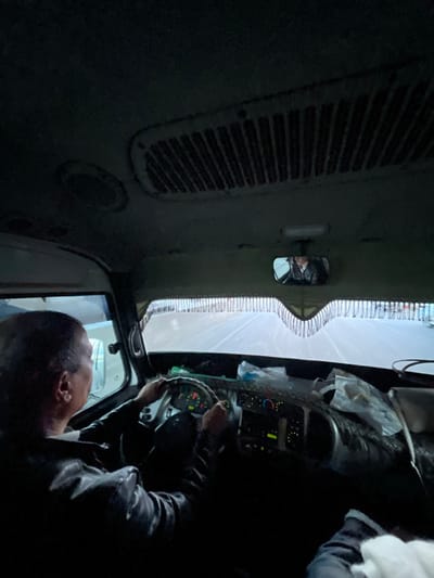 The interior of a vehicle, possibly a truck, is visible from an elevated perspective. A person is seated, presumably driving, with hands on the steering wheel and face visible in the lower right. The windshield is in the center, and a rearview mirror is on the left. The dashboard is visible, adorned with bags and other items. An air vent is on the left wall. The overall lighting is dim, contributing to a dark ambiance.