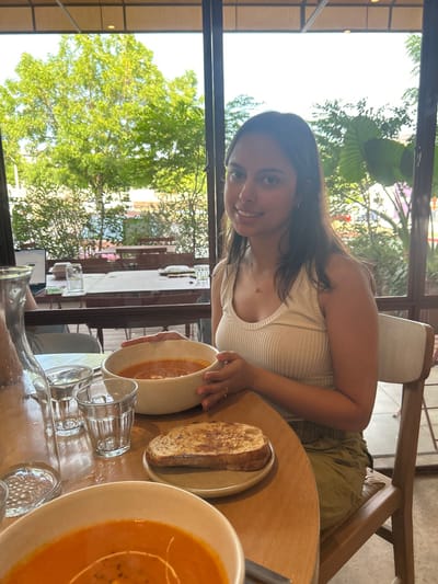 A woman in a white tank top sits at a round table laden with food. Two bowls of bright orange soup flank a small plate with grilled bread. Several small, empty glasses and a large water bottle are nearby. Sunlight streams through a window offering a glimpse of green trees outside, while other tables are visible in the background. She is looking towards the camera.