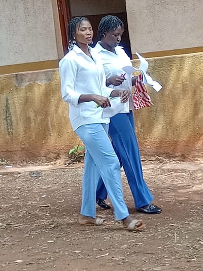 Two students stand outdoors in front of a building with a faded yellow facade. Both wear white, long-sleeved shirts. One has on blue pants and polished shoes, and is holding papers and a checkered cloth. The other student is wearing light blue pants and sandals, with a writing utensil in hand. The ground is bare earth with some small plants.