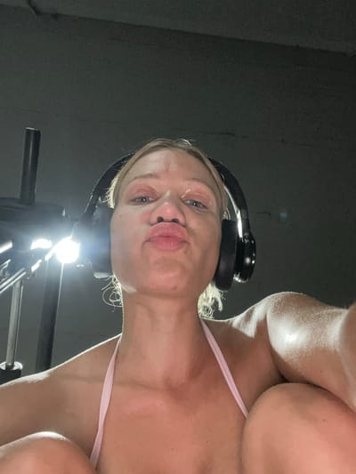 The image shows a woman with blonde hair wearing black headphones and a pink top. She is looking at the camera with her lips pursed. The lighting in the image is dim, with a bright light source in the foreground that appears to be a gym light. The background is dark and indistinct, suggesting an indoor setting such as a gym. There is no visible text in the image. The woman appears to be sweaty, which could indicate she has been exercising. The perspective of the photo is from below, looking up at the woman.