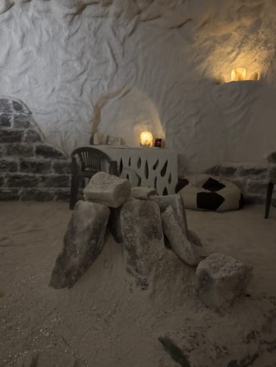 In the city of Korsuntsi, Ukraine, a salt cave relaxation room is depicted. The cave walls and ceiling are rough and white, made of salt. In the foreground, several large, irregularly shaped salt rocks are piled on a floor covered in fine, pale salt sand. To the left, a section of wall is constructed from rough-hewn, dark grey stones. In the background, a white counter with a decorative cutout pattern has a small digital clock and a glowing salt lamp on it. A dark plastic chair is positioned behind the counter. Further to the right, a large, round beanbag chair with a black and white checkered pattern rests on the salt floor. Two more salt lamps are visible, embedded in the salt wall to the right and further back in the cave, emitting a warm, yellow glow. The lighting is dim, with the salt lamps providing the primary illumination.