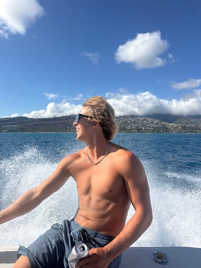 A single Caucasian male is seated on the white hull of a boat in motion on the ocean. He has blond hair, is shirtless, wears black sunglasses and a silver chain, and is clad in dark blue swim shorts. He is smiling and looking leftward and slightly upward, holding a crumpled silver beverage can in his right hand. A large volume of white water spray from the boat's movement is visible behind him. In the distant background, a mountain range covered with extensive residential development on its lower slopes is visible under a clear blue sky with scattered white clouds. The location is offshore from the coast of East Honolulu, Hawaii, United States.