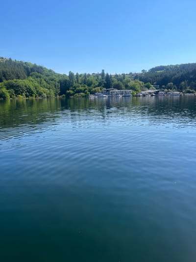 The image depicts a serene lake scene on a clear, sunny day. The foreground is dominated by the calm, blue water of the lake, with gentle ripples reflecting the sunlight. In the mid-ground, a lush, green forested hill rises from the opposite shore. A small cluster of buildings and several boats are visible along the shoreline, suggesting a harbor or a lakeside resort. The background is a vast expanse of clear blue sky. The location appears to be Brosh, Bulgaria, specifically a view of the lake with buildings and boats on the shore. There are no people or animals visible in the image. The scene is peaceful and tranquil, highlighting the natural beauty of the landscape and the man-made structures along the water.