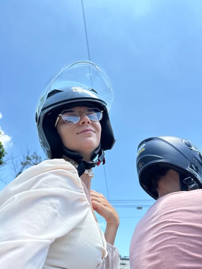 Two individuals wearing helmets are on a scooter, with a bright blue sky and some thin power lines visible in the background. The person in the foreground, positioned on the left, is a woman wearing round, gold-rimmed glasses and a white, flowy top. She is looking slightly upwards and to the right. A seashell necklace is visible around her neck. Behind her, to the right, is another person wearing a dark helmet and a pink t-shirt. The scene appears to be outdoors, possibly in a tropical or subtropical urban environment, given the clear sky and the presence of a scooter, which are common modes of transportation in cities like Hồng Sơn, Vietnam.