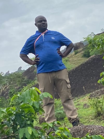 A man is standing outdoors in what appears to be a hilly, rocky landscape in Ganawuri, Nigeria. He is wearing a blue striped polo shirt, khaki pants, and sneakers. He has his hands on his hips, and he's looking toward the camera. The environment consists of rocks, vegetation, and a cloudy sky.