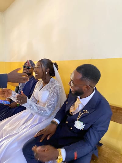 **Scene Description:**
**Location:** Interior setting, possibly a venue in Kuru, Nigeria.
**People:** A bride and groom are centrally located. The groom is wearing a dark blue suit, a gold-patterned tie, glasses, and a boutonniere. He has a gold watch on his left wrist. The bride is wearing a white, long-sleeved, heavily-embellished wedding dress and a veil. A person in a blue garment, wearing a striped hat and sunglasses, is partially visible to the left of the bride. Another person wearing a blue suit with their hand raised is also present in the lower left.
**Actions:** The bride and groom appear to be greeting someone or acknowledging congratulations. The groom is bending down slightly, and the bride is smiling and has her hand extended toward the other person with the outstretched arm.
**Interactions:** The primary interaction is between the bride and groom and a third individual. The two people seem to be reaching toward each other.
**Objects:**
* Wedding rings are visible on the bride's left hand.
* The grooms golden wristwatch is visible on his left wrist.
* Part of a mobile phone is visible in the bottom left
**Environment:**
* The backdrop is composed of cream and gold colored walls.
* The lighting seems bright, suggesting an interior space.