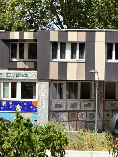 This is a vertical, medium shot of a modern building facade in Évry-Courcouronnes, France. The building features a striking design with alternating bands of dark gray and cream-colored panels. Interspersed among these panels are numerous windows with white frames. The facade is further embellished with colorful mosaic tile artwork.
To the left of the building, a large, leafy green tree with dense foliage partially obscures the view of the windows. To the right, the building's mosaic art becomes more prominent, featuring abstract patterns and depictions of celestial bodies, including planets, stars, and a rocket ship against a vibrant blue background. Some of the mosaic work appears to be in a grid pattern with square inserts. There is also a close-up view of vertical mosaic text that appears to read "ÉLYSÉES."
A single outdoor light fixture is mounted on a horizontal pole attached to the building. The scene is illuminated by natural daylight, suggesting it is daytime. The overall impression is one of an artistically designed, contemporary urban building with elements of nature and space-themed art.