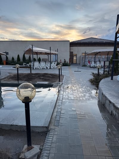 The image depicts an outdoor area in Kara-Oy, Kyrgyzstan. The scene appears to be adjacent to a building, possibly a resort or spa. Several white lounge chairs are arranged under awnings. A paved walkway leads past a shallow pool or water feature with spherical lampposts lining its edge. The walkway is wet, suggesting recent rain. The sky is cloudy with a hint of sunset.