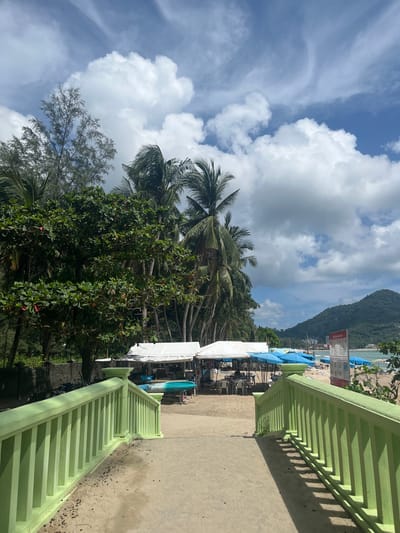 This is a photograph taken at a beach in Ban Bang Thao, Thailand. In the foreground, a light green railing is visible, leading down a concrete path. Beyond the railing, the sandy beach stretches out, dotted with white tents and numerous blue umbrellas, suggesting a market or food stalls. Palm trees and other lush green vegetation line the edge of the beach, and in the background, a verdant hill rises. The sky is a vibrant blue, with dramatic white clouds. A few people are visible under the tents, seated at tables. The overall impression is of a sunny day at a tropical beach resort.