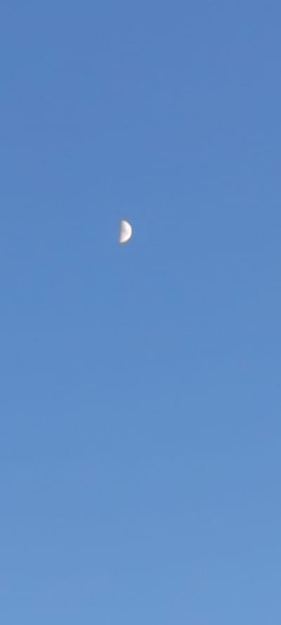 A single, partially illuminated moon is visible in a clear, bright blue sky. The moon appears as a crescent shape, with the illuminated portion being a lighter white and the shadowed portion a darker gray. The sky is a uniform color, with no clouds or other celestial bodies visible. The image has been captured during daylight hours, as evidenced by the clear blue sky. The location is Supsekh, Russia.