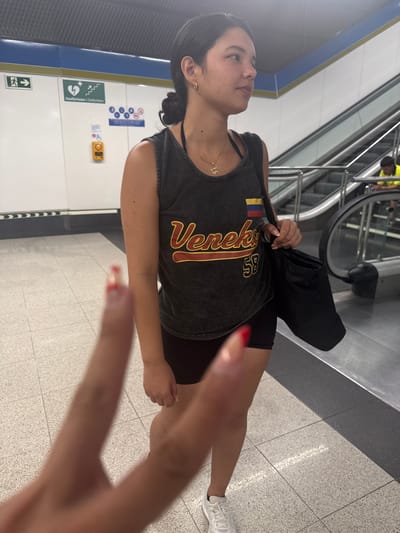The image shows a young woman standing in a subway station. She is wearing a dark tank top with "Veneko 58" printed on it and black shorts. She has a black tote bag slung over her shoulder and is looking to her right. A blurry finger in the foreground is positioned to look like it is pointing at her. In the background, an escalator is visible, with another person in a yellow vest at the bottom. The walls of the station are white with blue and yellow trim, and there are signs indicating emergency exits and a defibrillator. The floor is tiled with a light gray and white speckled pattern. The image appears to have been taken in Getafe, Spain.
