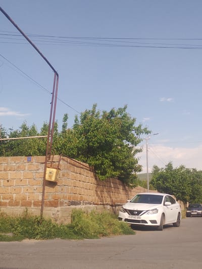 The image depicts a street scene in the city of Nor Geghi, Armenia. In the foreground, a white Nissan sedan is parked on the right side of the asphalt road, facing towards the viewer. The car has its license plate visible, reading "37 KE 695". To the right of the Nissan, a dark-colored Mercedes-Benz sedan is parked further down the road, also facing towards the viewer.

A brick wall, constructed from yellowish-tan bricks, runs along the left side of the road. A rusted metal pole stands adjacent to the wall, with a yellow electrical box attached to it. A blue square sign with the number "16" is visible on the brick wall, above the electrical box. Green grass and vegetation grow along the base of the wall.

Behind the wall, lush green trees with dense foliage dominate the mid-ground. In the background, a clear blue sky with scattered white clouds is visible, along with the silhouette of a distant hill. Overhead, power lines stretch across the sky, supported by poles. The overall scene suggests a quiet, residential area on a sunny day.