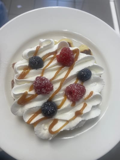A top-down view captures a dessert served on a white plate. The dessert features several small pancakes topped with a generous swirl of whipped cream. Drizzled over the whipped cream are thin lines of caramel sauce. Scattered atop the cream are fresh raspberries and blueberries, some dusted with what appears to be powdered sugar. The overall presentation suggests a sweet and indulgent treat. The setting appears to be indoors, possibly a restaurant or kitchen, given the clean white plate and the indistinct background. The lighting is bright and even, indicating daytime or well-lit interior. No people are visible in the frame, and no text is present. The focus is solely on the appetizing dessert. The dessert is a single serving, presented neatly on a round white plate. The whipped cream is piped in decorative peaks, creating a visually appealing texture. The caramel drizzle adds a golden contrast to the white cream and red and blue berries.