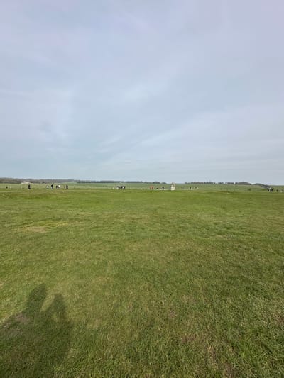The image displays the ancient monument of Stonehenge, located approximately 2 miles west of Amesbury, Wiltshire, United Kingdom. The scene is an expansive, open grassy plain under an overcast, light grey sky. In the middle distance, slightly right of center, the large, grey standing stones of Stonehenge are visible. A low, light-colored barrier or rope fence encircles the immediate perimeter of the monument. Numerous individuals, appearing as small, dark figures, are scattered across the field, primarily observing the monument from a distance or walking along faint pathways. The foreground consists of green, somewhat uneven grass. The shadows of three indistinct human figures are cast onto the grass in the bottom-left corner of the frame, originating from outside the image to the lower left. The horizon line is flat, with faint treelines and distant structures visible.