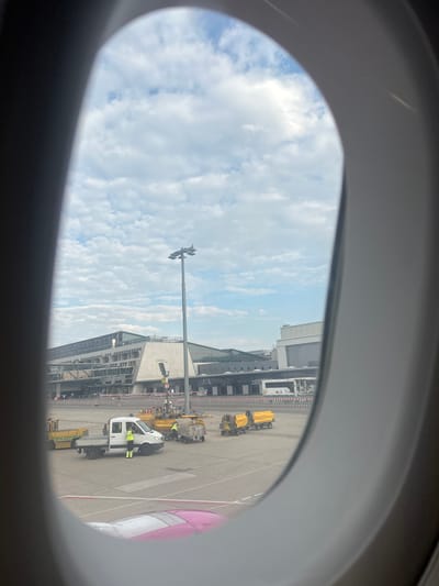 This image, taken from an airplane window, shows the exterior of a modern airport terminal with a cloudy sky overhead. In the foreground, the wing of a pink and white airplane is visible. The tarmac is populated with various service vehicles, including a white van with a flatbed, two yellow carts, and other yellow ground support equipment. Several individuals in bright yellow safety vests are visible near the vehicles, suggesting airport operations are in progress. The airport building features a distinctive angular architectural style with extensive glass facades. The scene is consistent with Stuttgart Airport (STR), located in Echterdingen, Germany.