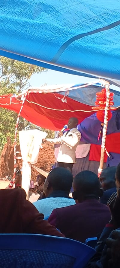 The image depicts a speaker addressing a crowd under a makeshift canopy, possibly in an outdoor setting in Kalemie, Democratic Republic of the Congo. The canopy is made of blue and red tarps supported by wooden posts. The speaker, wearing a light-colored suit, is holding a microphone and gesturing with his right hand. The audience, seated in blue plastic chairs, appears to be attentive. The environment includes trees and other vegetation in the background, suggesting a rural or semi-rural location. Additional people are sitting on the ground behind the speaker. Red decorative tassels are visible near one of the support poles. The overall impression is that of a public gathering or event.